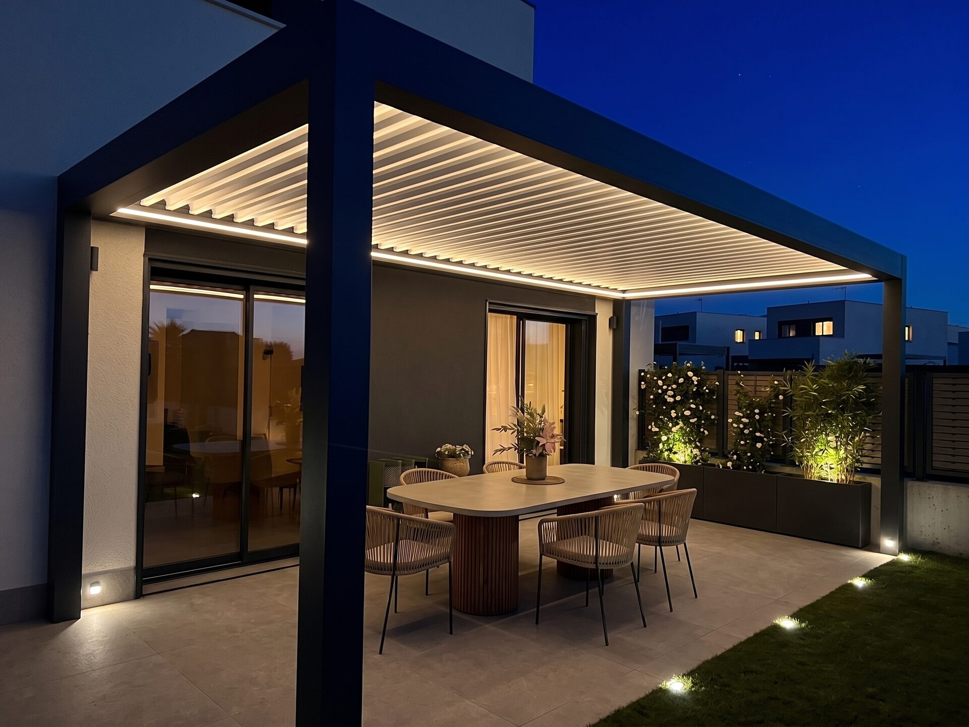 Pérgola bioclimática CERCA con lamas abiertas sobre una terraza con mesa de comedor para ocho y jardín con seto, iluminación LED integrada al anochecer